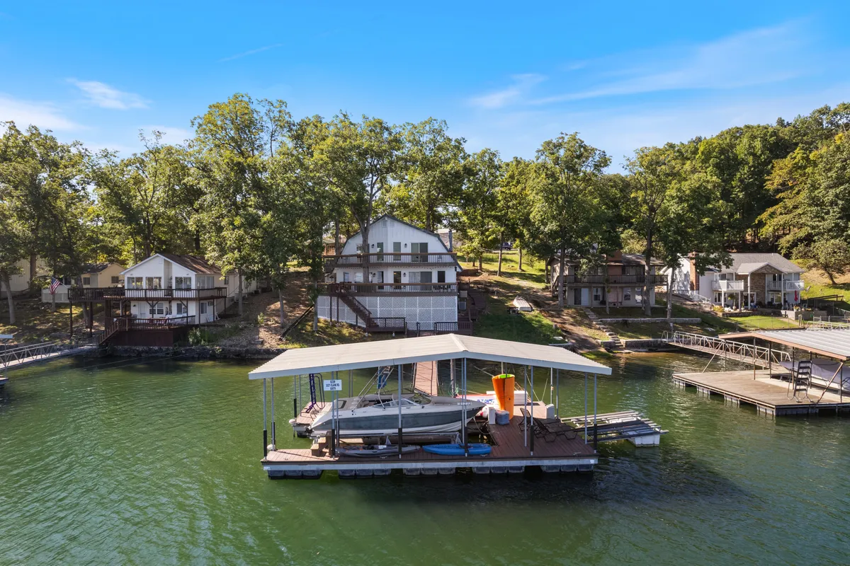 A picturesque lakefront scene featuring houses, a boat dock, lush greenery, and a clear blue sky. The inviting dock and natural setting highlight the ease of gathering for swimming, fishing, or enjoying evening bonfires by the water.