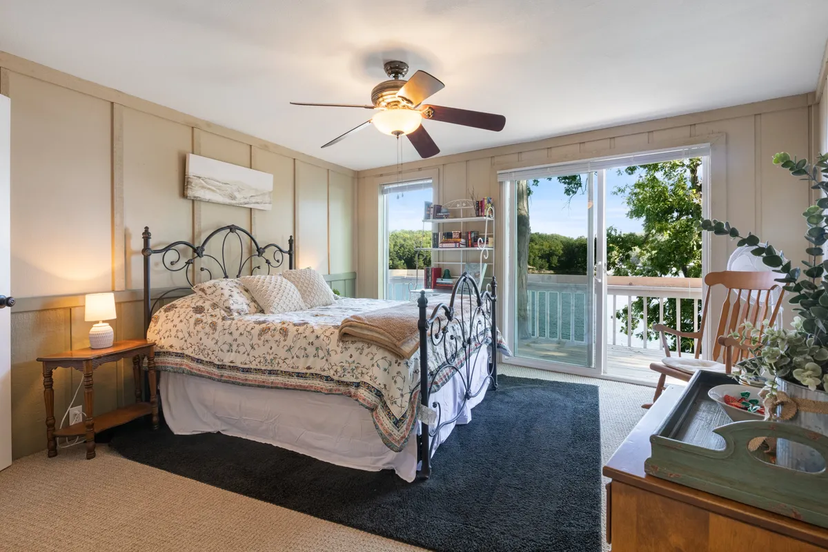 A cozy bedroom with a decorative bed, soft bedding, and a large window offering a scenic view of the lake. The corner features a tidy bookshelf, suggesting a tranquil place for reading or relaxing. Sunlight streams in, creating a warm, peaceful atmosphere.
