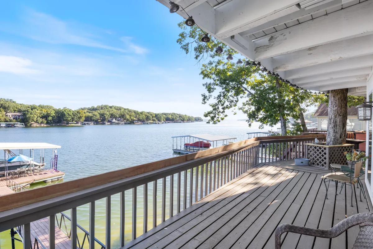 A spacious deck stretches out over the lakeshore, offering sweeping views of the lake and distant boathouses, framed by lush greenery. The inviting outdoor space is perfect for sipping morning coffee or enjoying peaceful evenings.