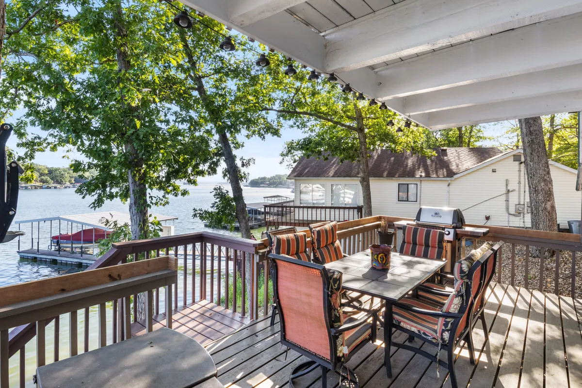 A spacious outdoor dining set is arranged on a wooden deck overlooking a sparkling lake, with the house and surrounding trees providing a tranquil, scenic backdrop. The setting is ideal for al fresco meals, with direct access to the water for swimming or launching kayaks.