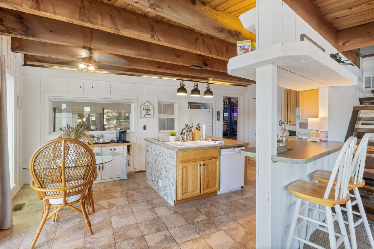 A bright, open kitchen and dining area with a large island, wooden beams overhead, and shelves filled with decor. The dining area invites gatherings, and the setup is perfect for meal prep or casual conversation, with plenty of light coming in from the windows.