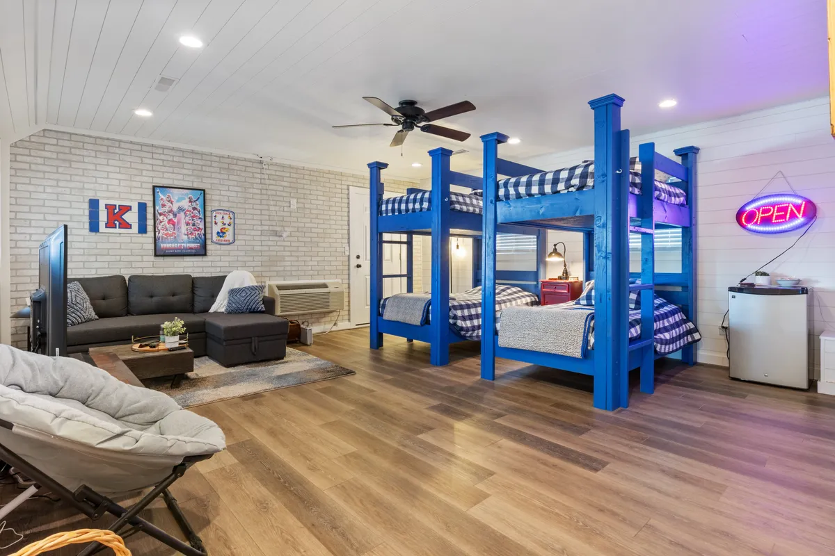 This image features a vibrant bunk room with bunk beds, a comfortable sofa, and playful decor like a neon 'OPEN' sign and wall artwork. The setting is designed for both sleeping and socializing, capturing the essence of a family- or group-friendly space where kids and adults can gather, play games, or relax.