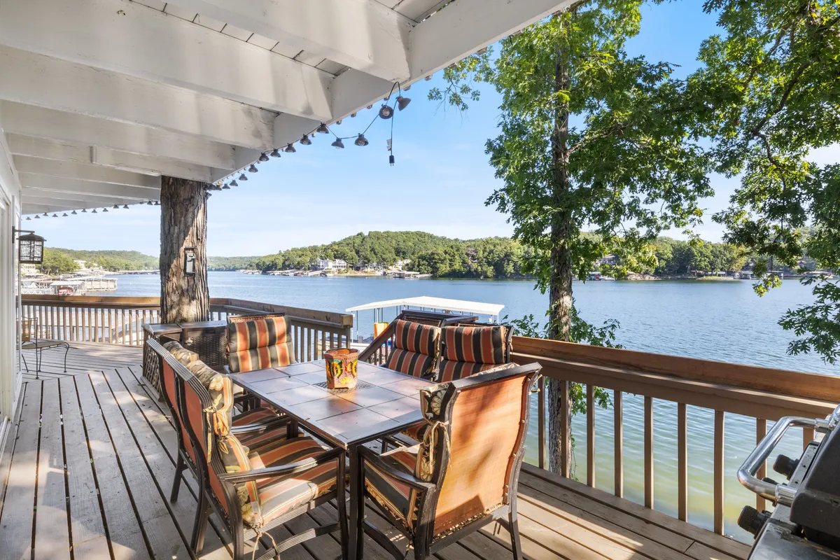An outdoor dining table with comfortable chairs sits under the open sky, overlooking the serene lake. Decorative lanterns and lush green trees create a warm, friendly setting for al fresco meals and social gatherings.