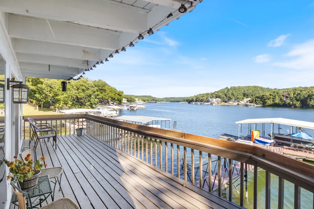 A serene lakeside deck with wooden rails overlooks calm water, dotted with boats and surrounded by lush green trees. Decorative string lights hang above, adding a romantic touch for relaxing or socializing at sunset.