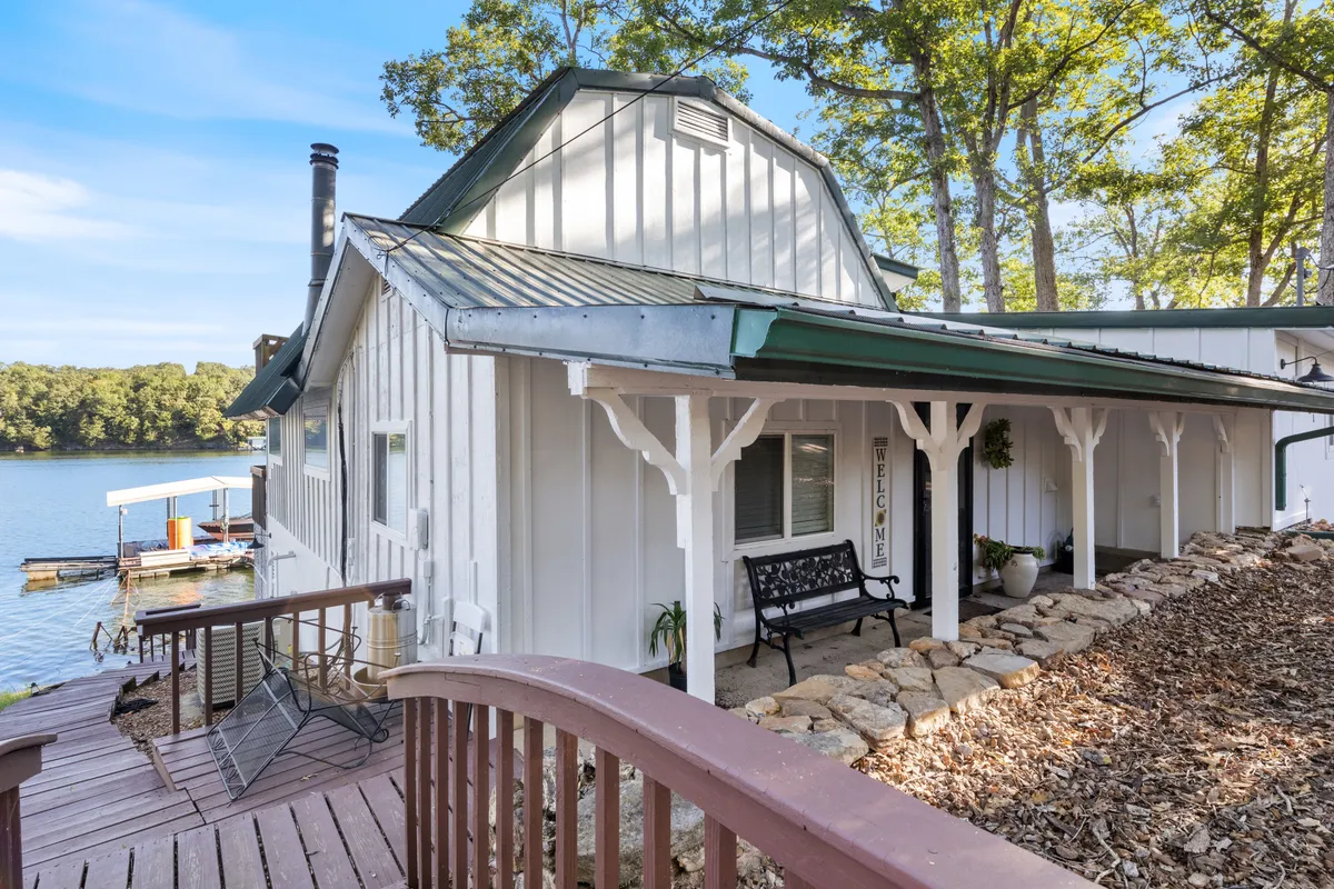 A welcoming lakeside porch with a bench, wooden deck, and a friendly sign faces the shimmering lake, providing a cozy spot to relax, watch sunsets, or share stories. The tranquil water and greenery set a peaceful, romantic scene.