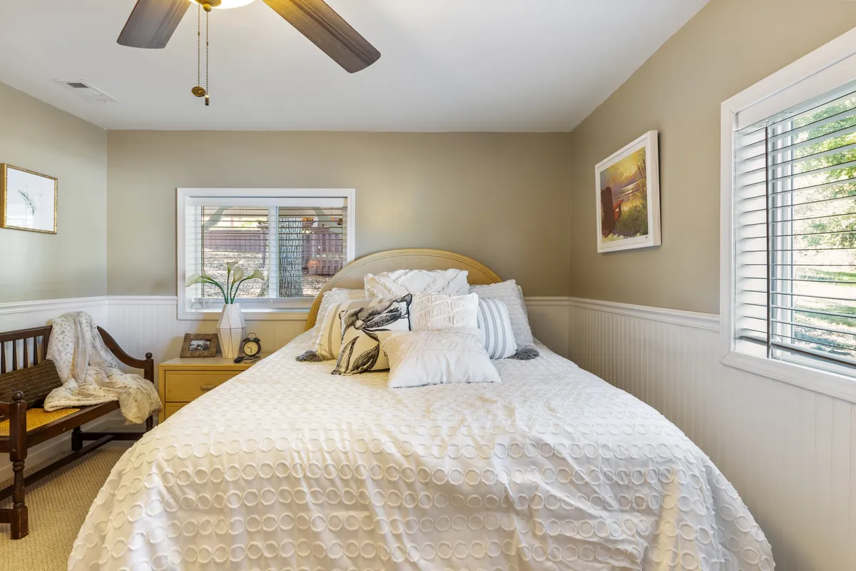 A serene bedroom featuring a bed adorned with decorative pillows, a window that allows sunlight to gently filter in, and a wooden chair draped with a cozy blanket. The overall mood is calm and inviting, perfect for peaceful retreats.