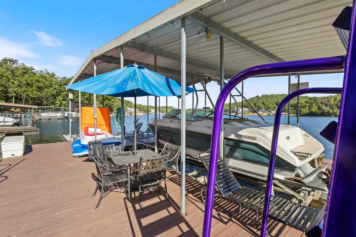A welcoming lakeside boathouse scene with boats, a shaded dining table, umbrella, and water views. Friends can be imagined enjoying sunset dinners, relaxed conversation, and fun-filled days on the water.