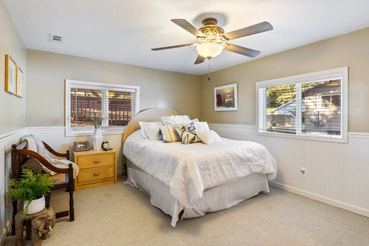 A bright, soothing bedroom with crisp white bedding, a ceiling fan, and a wooden chair beside the window. A nightstand with a lamp adds a homely touch, while natural light fills the space, creating a restful retreat.