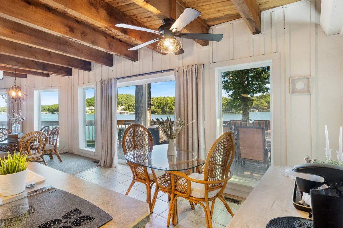 A spacious dining table set beside large windows, allowing ample natural light to fill the room. Wooden beams and a ceiling fan add to the cozy, communal feel, with views out to the lake visible through the windows.
